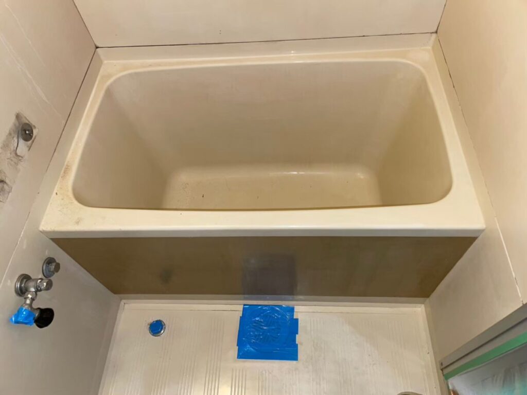 神戸市灘区の施工前、ユニットバスの浴槽と茶色っぽいエプロン。床に青いテープが貼られている
