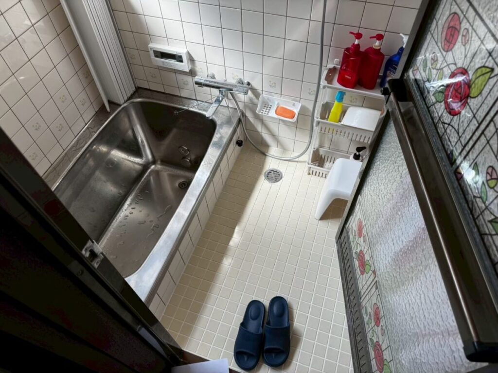 施工前、浴室を入口側から見たところ。左にステンレス浴槽、床はクリーム色の小さなタイル、右に濃い茶色の枠のドア