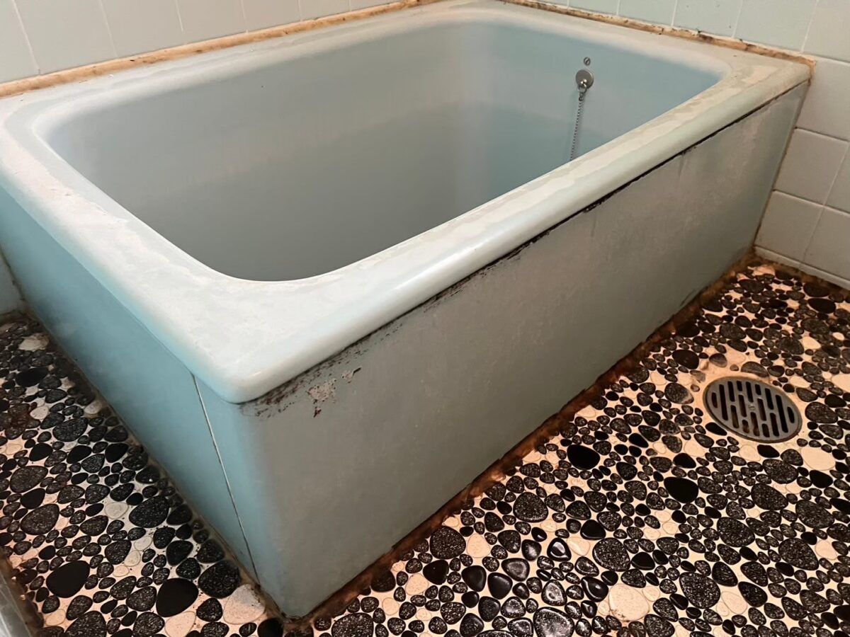 施工前、水色のホーロー浴槽を斜めから見たところ。ふちに白っぽい汚れ、前面の上端に黒っぽい汚れがある