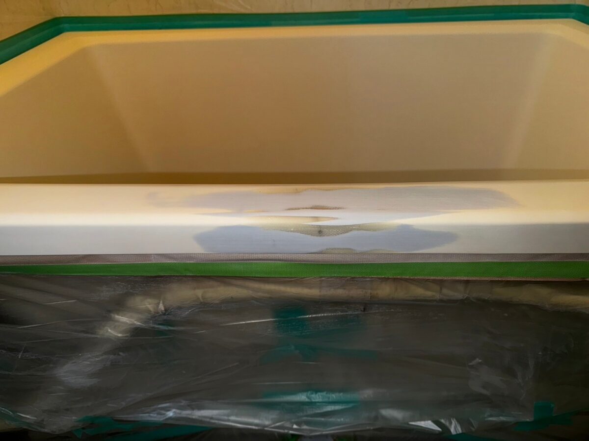 施工の途中、浴槽のふちの正面。灰青色と白っぽい部分がまだら状に広がり、下は緑のテープとビニールで養生されている