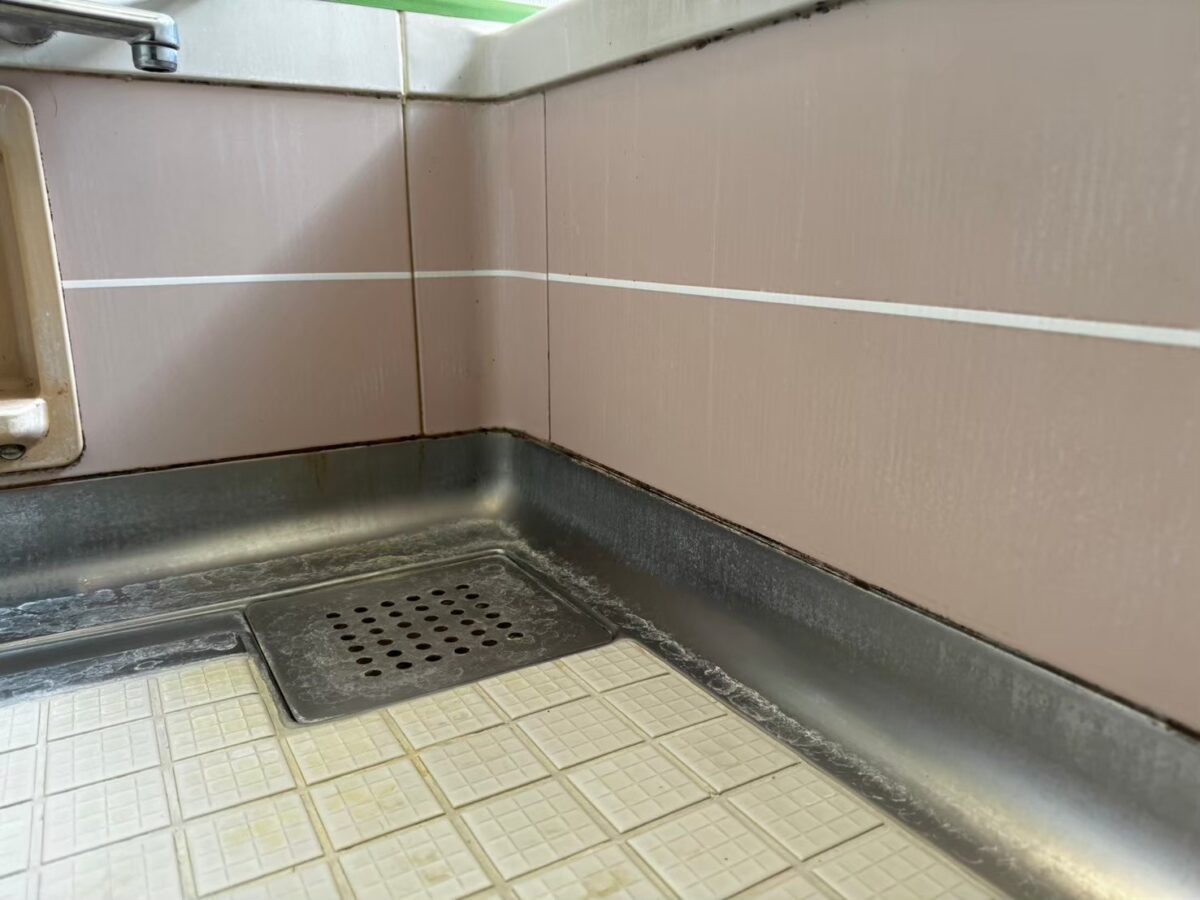 施工前、洗い場の隅。排水口の金具、ピンク色の壁、金属の立ち上がり。床は白っぽい小さな四角が並ぶ柄